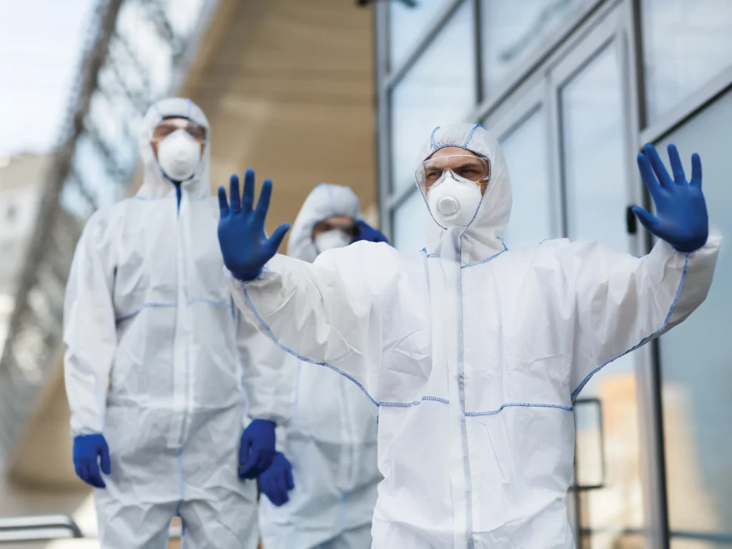 A person wearing a medical mask, gloves and protective suit holds their hands up as a signal to stop.