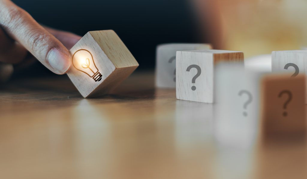 Five small wooden blocks sit on a table. Four of the blocks have question marks drawn on them, and one has a lit-up lightbulb drawn on it. Fingers place the block with the lightbulb in front of the blocks with question marks on them.