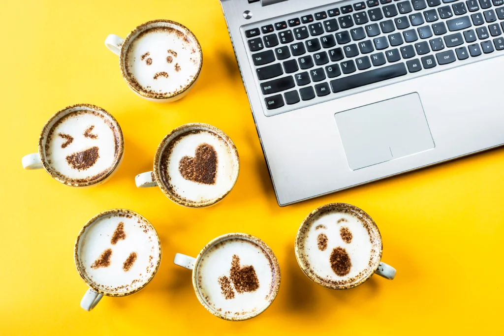 Cappuccinos sit beside an open laptop. The chocolate powder on top of each coffee forms an emojis sad face, laughing face, heart, angry face, thumbs up and surprised face.